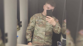 Un joven viste un uniforme militar de camuflaje de Estados Unidos y sostiene un teléfono inteligente frente a un espejo