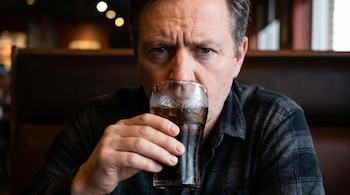 Primer plano de un hombre caucásico de mediana edad, con ceño fruncido y expresión seria, bebiendo de un vaso de refresco con hielo, vestido con una camisa a cuadros.
