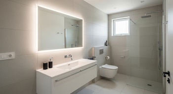 Interior de un baño con azulejos claros, lavabo blanco, espejo rectangular iluminado con LED, inodoro suspendido y ducha con mampara de cristal.
