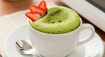 Primer plano de un bizcocho de té verde en una taza blanca, espolvoreado con matcha y decorado con dos rodajas de fresa, sobre un platillo blanco con una cuchara.