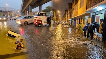 Una calle inundada por la lluvia muestra vehículos sumergidos, personas colocando sacos de arena y un hombre con un niño en brazos.