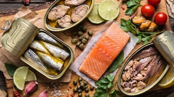 Filete de salmón, latas abiertas de sardinas y mariscos, rodajas de lima, tomates cherry y alcaparras sobre una tabla de madera