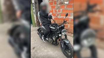 Un hombre con el rostro pixelado viste chaleco negro y está sentado sobre una motocicleta negra frente a una pared de ladrillos vistos