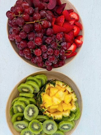 Dos cuencos de madera con uvas rojas, fresas en mitades, rodajas de kiwi, piña en cubos y piezas de fruta roja, sobre una superficie blanca