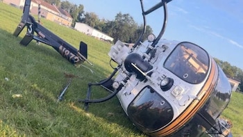 Un helicóptero blanco y negro volcado en un área de hierba. El fuselaje está de lado, con la cola hacia arriba. Se aprecian árboles y casas detrás