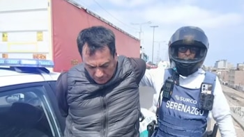 Un hombre esposado es llevado por un agente de seguridad con casco y chaleco de Surco Serenazgo, junto a un coche policial y un camión en una carretera