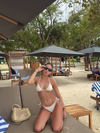 Una mujer en bikini blanco y lentes de sol posa sentada en un camastro de playa, junto a un bolso tejido. Fondo con arena, árboles y quitasoles