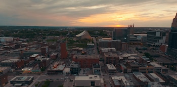Vista aérea de una ciudad al atardecer, con edificios de diferentes alturas, calles transitadas por vehículos y un cielo con nubes anaranjadas