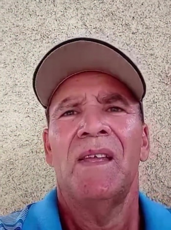 Primer plano de un hombre maduro de piel clara con gorra beige y polo azul, mirando directamente a la cámara sobre un fondo texturizado