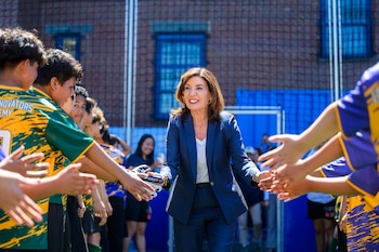 La gobernadora Kathy Hochul, sonriendo, choca las manos con varios niños en uniformes deportivos en una cancha exterior