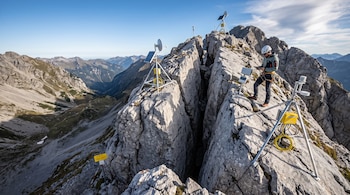 Un hombre con casco trabaja junto a una grieta profunda en la cumbre rocosa de una montaña rodeada de equipos de medición y paneles solares.
