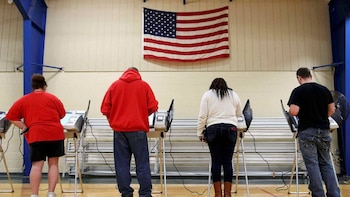 Cuatro personas votan en cabinas electorales individuales con una bandera de Estados Unidos colgada en la pared. El suelo es de madera y las paredes claras