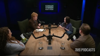 Conan O'Brien y Billie Eilish sentados frente a frente en una mesa de madera con micrófonos, conversando en un estudio de podcast con iluminación azul