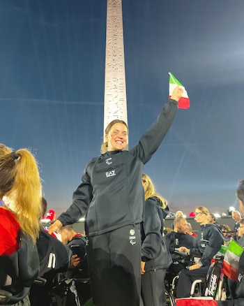 Una mujer con uniforme deportivo de Italia sostiene una pequeña bandera italiana frente a un obelisco iluminado de noche