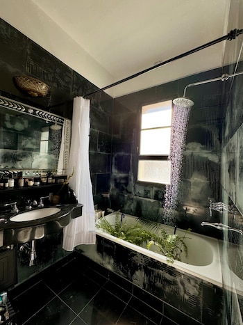 Baño con paredes y suelo de azulejos negros, lavabo con espejo, cortina blanca, bañera con plantas y ducha de lluvia en funcionamiento