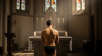 Hombre de espaldas en una iglesia mirando al altar, con el torso descubierto y ropa interior negra. Altar con crucifijo, cuatro velas encendidas, y vitrales.