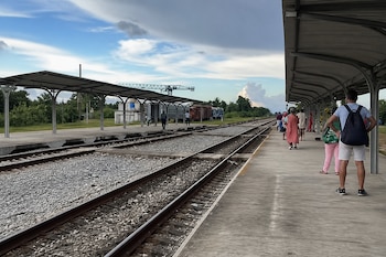 Varios pasajeros permanecen en un andén techado junto a dos vías de tren con balasto y vagones al fondo.