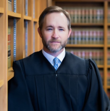 Hombre con barba y pelo corto usando una toga judicial negra y corbata azul a rayas. Fondo con estanterías de madera llenas de libros