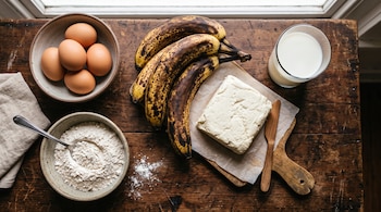 Plátanos maduros, un bloque de queso sobre una tabla, huevos, harina en un cuenco y un vaso de leche sobre una mesa de madera.
