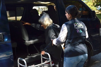 Una mujer con cabello blanco y andador sube a una Minivan oscura, siendo asistida por una mujer con chaleco de la PGN