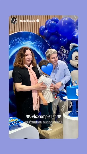Lizy Tagliani y un hombre posan junto a un niño con el rostro cubierto, frente a una torta de cumpleaños y globos azules