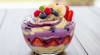 Cuenco de vidrio con capas de crema amarilla y morada. Incluye fresas, arándanos, frambuesas, plátano en rodajas, semillas de chía y hoja de menta sobre mesa de madera.