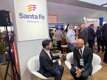 Dos hombres con traje conversan sentados en sillones blancos frente a un cartel con el texto Santa Fe Provincia en una exposición