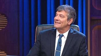 Un hombre de mediana edad, con cabello gris, traje oscuro y corbata azul a puntos, sonríe y mira hacia la derecha, sentado en una silla negra