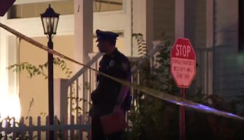 Un oficial de policía, cinta policial amarilla, un farol, un cartel de stop rojo, una valla blanca, arbustos y la fachada de una casa de noche