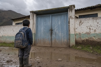Niño de espaldas con mochila y botas en un camino con barro frente a un portón de madera azul cerrado y paredes deterioradas.