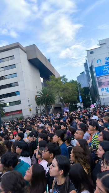 Una multitud de personas reunida en una calle urbana, con edificios de oficinas, árboles y cielo con nubes de fondo