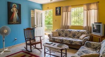 Sala con paredes azules y amarillas, dos sofás con estampado floral, mecedora, mesa de centro, televisor de tubo, ventilador de pie y dos cuadros religiosos.