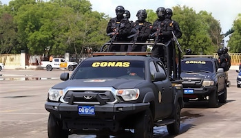 Agentes con uniforme negro y armas largas permanecen de pie en la plataforma de una camioneta negra con la inscripción CONAS