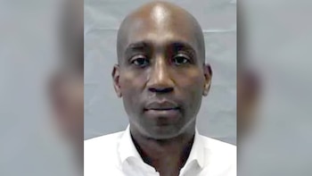 Un hombre de piel oscura y cabeza rapada viste una camisa blanca con cuello abierto sobre un fondo gris claro
