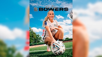 Mujer rubia con cola de caballo vestida con camiseta y pantalones cortos de fútbol, agachada en césped verde junto a un balón. Cielo azul con nubes blancas