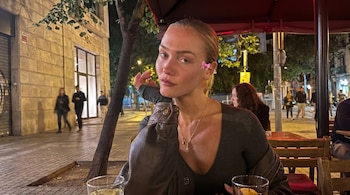 Mujer rubia con lazo rosa en la oreja sentada en la terraza de un café por la noche. Viste chaqueta oscura y top V. Al fondo, calle urbana iluminada con personas.