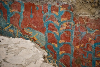 Mural antiguo en tonos rojos, azules y amarillos, mostrando una lagartija de cuerpo azul y amarillo, con plantas estilizadas de maíz y elementos abstractos