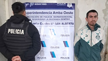 Un hombre detenido, con sudadera verde y blanca, posa junto a un oficial de policía de espaldas, frente a un cartel azul de la Policía de Buenos Aires