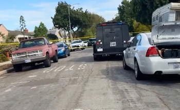 Un vehículo blindado policial, automóviles y una patrulla con el maletero abierto en una calle residencial acordonada con cinta amarilla