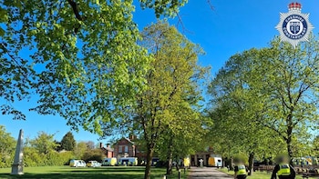 Vista exterior de una propiedad con vehículos policiales y agentes en un día soleado, rodeada de árboles verdes y un monumento obelisco blanco en Crewe