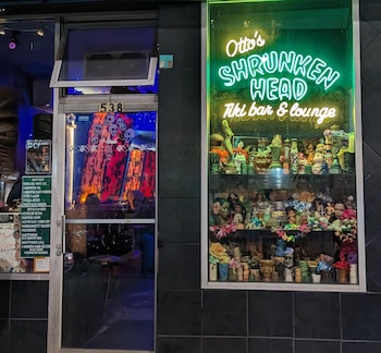 Fachada de bar con letrero de neón verde 'Otto's Shrunken Head', vitrina con figuras tiki, muñecas y flores, y puerta de vidrio con calaveras y número 538