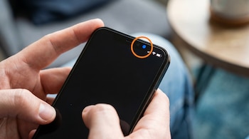 Primer plano de manos sosteniendo un celular con pantalla oscura. En la esquina superior derecha de la pantalla se ve un punto azul, enmarcado por un círculo naranja.