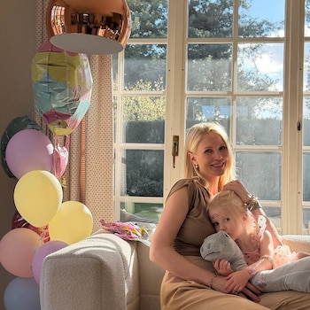 Daniela Christiansson, con una blusa marrón, abraza a su hija Elle, vestida de rosa, sentadas en un sofá con globos de colores detrás y una ventana