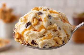 Una cuchara metálica con una porción de helado blanco, jarabe de caramelo, trozos de chocolate oscuro y castañas de cajú.