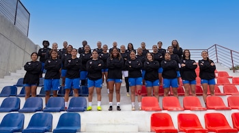 Un grupo de aproximadamente treinta mujeres jóvenes vestidas con sudaderas negras y pantalones cortos azules posa en las gradas de un estadio.