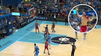 Una vista aérea de una cancha de baloncesto con varios jugadores y un árbitro; en un círculo superpuesto a la derecha, un jugador sin camiseta celebra con brazos levantados.