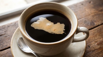 Vista superior de una taza de café oscuro con la silueta espumosa del mapa de El Salvador. La taza está sobre un plato y una cuchara en una mesa de madera.