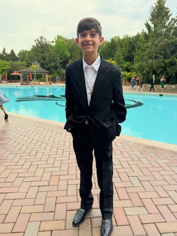 Un niño vestido con traje negro, camisa blanca y medalla dorada sonríe de pie en un patio adoquinado junto a una piscina