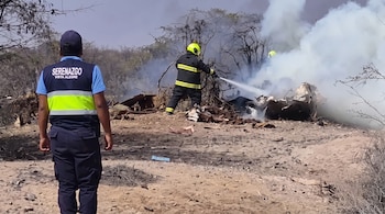 Una persona con gorra y chaleco de Serenazgo Vista Alegre observa a un bombero que extingue un incendio en los escombros de un avión en un terreno árido