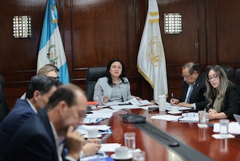 Una mujer en mesa de reuniones con varias personas, documentos y tazas; banderas de Guatemala y del Ministerio de Finanzas Públicas al fondo